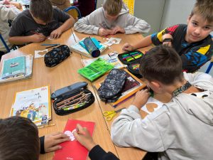 Grupa chłopców siedzi przy szkolnym stoliku i pracuje nad wycinankami w kształcie serca. Na biurku leżą książki, zeszyty, piórniki i nożyczki. Jeden z chłopców pisze coś różowym ołówkiem na papierowym sercu, a inny wycina jego kształt.