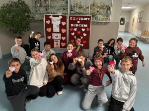 Grupa dzieci pozuje wspólnie w szkolnym korytarzu przed gazetką ścienną zatytułowaną „Czytamy z sercem”. Każde dziecko trzyma w ręku małe papierowe serce z napisem. W tle widać gablotę z pucharami i trofeami sportowymi.