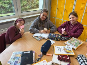 Trzy dziewczynki siedzą przy szkolnym stoliku i uśmiechają się, trzymając w dłoniach różowe i białe papierowe serca. Na stole leżą zeszyty ćwiczeń, książki, piórniki i przybory szkolne. Za nimi stoją żółte szafki uczniowskie.