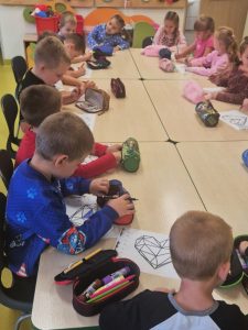 Dzieci siedzą przy połączonych stolikach w sali szkolnej i kolorują obrazki w kształcie geometrycznego serca. Na stołach leżą przybory szkolne w piórnikach. Większość dzieci jest pochylona nad rysunkami i skupiona na pracy.