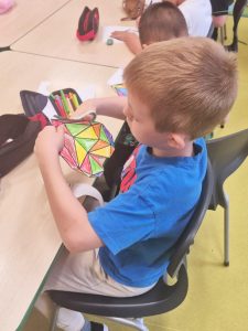 Chłopiec siedzi przy stoliku i trzyma w ręku wycięty obrazek geometrycznego serca pokolorowanego w różne barwy. Na stole leży piórnik i przybory szkolne, a w tle widać inne dzieci zajęte rysowaniem i kolorowaniem.