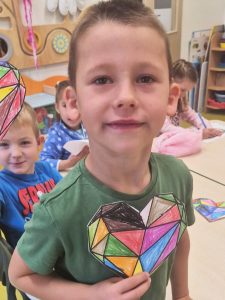 Chłopiec w zielonej koszulce stoi w sali szkolnej i trzyma przed sobą pokolorowany obrazek w kształcie geometrycznego serca w wielu barwach, m.in. czarnym, zielonym, różowym i niebieskim. W tle widać inne dzieci siedzące przy stoliku i rysujące.