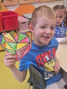 Chłopiec w niebieskiej koszulce z postacią SpongeBoba siedzi przy szkolnym stoliku i uśmiecha się, pokazując pokolorowane geometryczne serce w odcieniach zieleni, czerwieni i żółci. Za nim siedzą inne dzieci pracujące nad rysunkami.