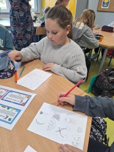 Dziewczynka w szarej bluzie siedzi przy szkolnym stoliku i rysuje na kartce. Na stole leży kilka kartek z rysunkami warzyw i owoców, m.in. marchewki, banana, arbuza, czereśni i truskawek. Obok znajdują się piórniki z kredkami i ołówkami.