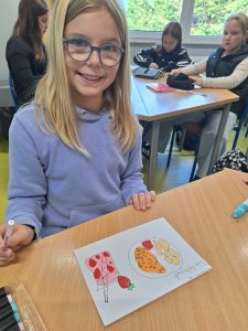 Dziewczynka w okularach i fioletowej bluzie siedzi przy szkolnym stoliku i uśmiecha się do kamery. Na biurku leży kartka z kolorowym rysunkiem przedstawiającym loda, truskawkę oraz talerz z jedzeniem.