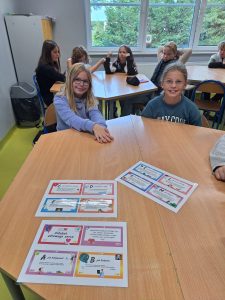 Dzieci siedzą przy stoliku w klasie. Na stole rozłożone są kolorowe plansze edukacyjne zatytułowane „Alfabet zdrowego serca”. W tle widać inne dzieci przy sąsiednich stołach.