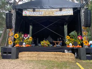Zdjęcie udekorowanej przez pracowników szkoły sceny. Nad sceną rozwieszony jest baner z napisem: "DOŻYNKI GMINA ŁYSOMICE".