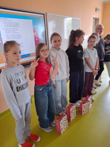 Grupa dziewczynek stoi w szeregu w klasie przed tablicą multimedialną. Każda z nich ma przed sobą kolorową torbę upominkową. Jedna dziewczynka pokazuje znak „V” palcami w kierunku aparatu.