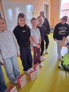 Siedem dziewczynek stoi obok siebie w klasie przy tablicy. Przed nimi ustawione są kolorowe torby z upominkami. Wszystkie patrzą w stronę fotografa i uśmiechają się.