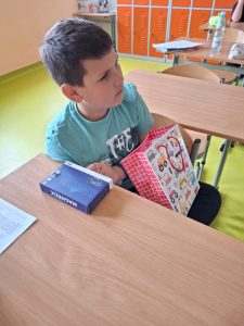 Chłopiec siedzi w szkolnej ławce, trzyma w ręku kolorową torbę w prezencie. Na biurku obok leży pudełko z napisem „Magnetic” oraz kartka z tekstem.