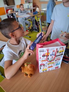 Chłopiec w okularach siedzi w szkolnej ławce, trzyma pudełko z napisem „Magnetic” i rozmawia z kolegą. Na stole obok leży maskotka i torba upominkowa w kolorowe pojazdy.