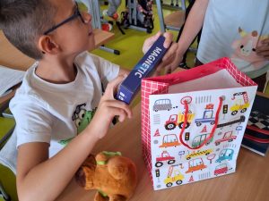 Chłopiec siedzi w szkolnej ławce i trzyma w rękach pudełko gry Magnetic – prezentu z okazji Dnia Chłopaka. Obok na stole stoi torba upominkowa w kolorowe pojazdy.