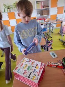 Chłopiec w niebieskiej bluzie z napisem „Lemon Explore” stoi przy ławce i ogląda pudełko z napisem „Magnetic”. Na stole leży torba upominkowa i baton czekoladowy.
