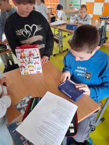 Chłopiec w niebieskiej bluzie siedzi przy szkolnej ławce i trzyma w ręku pudełko z napisem „Magnetic”. Obok niego stoi dziewczynka z torbą upominkową w kolorowe pojazdy.