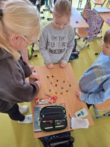 Troje dzieci pochyla się nad ławką, rozgrywając grę Magnetic podarowaną chłopcom na Dzień Chłopaka. Wewnątrz sznurka ułożone są czarne magnetyczne elementy, obok leżą przybory szkolne i baton.