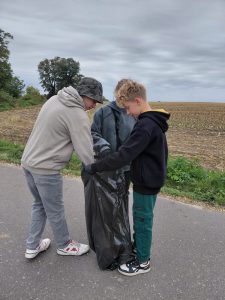Troje uczniów klasy siódmej stoi na asfaltowej drodze wokół trzymanego przez nich worka na śmieci.