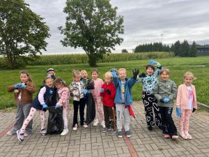 Uczniowie z oddziału przedszkolnego pozują do wspólnego zdjęcia na parkingu przed szkołą. Dzieci mają założone na dłoniach gumowe rękawiczki. Dwie dziewczynki trzymają przed sobą duży worek na śmieci.