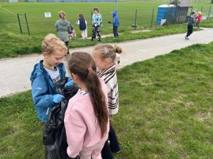 Uczniowie zbierają śmieci na terenach otaczających szkołę. Na pierwszym planie widoczne są dwie dziewczynki podchodzące do chłopca, który trzyma przed sobą worek na śmieci.