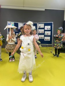 Grupa dzieci w szkolnej sali prezentuje stroje z recyklingu. Na pierwszym planie dziewczynka w białym stroju z plastikowych odpadów. W tle stoją inne dzieci w strojach ekologicznych, m.in. dziewczynka z parasolem z papieru i spódnicą z rolek tekturowych oraz chłopiec ubrany w strój z czarnego worka na śmieci ozdobionego zielonymi i pomarańczowymi elementami oraz kartonowymi kółkami, który na głowie ma czarny kapelusz udekorowany kolorowymi nakrętkami.