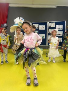 Grupa dzieci w szkolnej sali prezentuje stroje z recyklingu. Na pierwszym planie dziewczynka ubrana w sukienkę wykonaną z kolorowych worków na śmieci.. W tle stoją inne dzieci w strojach ekologicznych, m.in. pirat z opaską na oku, dziewczynka z parasolem z papieru i spódnicą z rolek tekturowych oraz dziewczynka w białym stroju z plastikowych odpadów.
