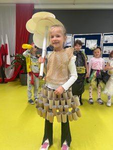 Grupa dzieci bierze udział w szkolnym wydarzeniu ekologicznym. Na pierwszym planie stoi dziewczynka ubrana w strój wykonany z papierowych rolek po papierze toaletowym. Dziewczynka trzyma w dłoni parasol wykonany z papierowych tubek i talerzy. W tle widać inne dzieci w strojach z materiałów recyklingowych, m.in. chłopca w stroju pirata oraz inne dzieci w białych i kolorowych kreacjach z odpadów.