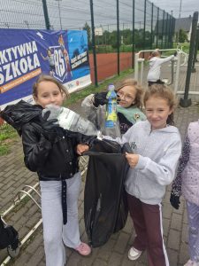 Trzy uczennice wkładają plastikowe butelki do worka naśmieci, który wspólnie trzymają. Dziewczynki stoją na boisku.