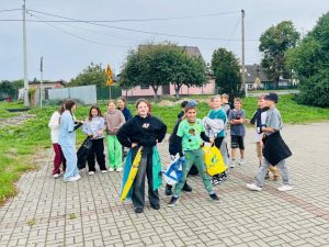 Uczniowie klasy piątej stoją na parkingu przed szkołą. Dzieci trzymają w dłoniach worki na śmieci. Na pierwszym planie widoczna jest dwójka uczniów prezentująca swoje stroje wykonane z materiałów z recyklingu.