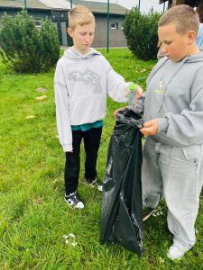 Dwóch chłopców stoi na trawniku. Jeden z nich trzyma worek na śmieci, a drugi wrzuca do niego plastikową butelkę.