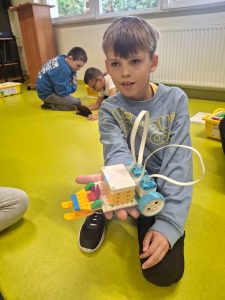 Chłopiec w niebieskiej bluzie klęczy na jasnozielonej podłodze w klasie i pokazuje do kamery zbudowany z klocków LEGO robotyczny pojazd z kołami, czujnikami i kolorowymi elementami. W tle dwójka innych dzieci siedzi na podłodze i patrzy w tablet.