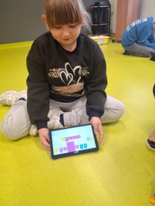 Dziewczynka w czarnej bluzie z nadrukiem siedzi na jasnozielonej podłodze i trzyma tablet. Na ekranie widać kolorowe bloczki programu w aplikacji do nauki kodowania, ułożone w sekwencję poleceń. W tle dwójka dzieci pracuje nad klockami.