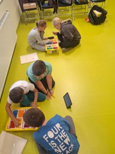 Czwórka dzieci siedzi na jasnozielonej podłodze w sali i pracuje w dwóch grupach. Każda grupa ma przed sobą zestaw klocków edukacyjnych LEGO Education w żółtym pudełku. Obok dzieci znajduje się tablet, na którym prawdopodobnie wyświetlane są instrukcje. W tle widać krzesła oraz plecaki.