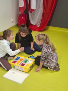 trzy dziewczynki siedzą na podłodze obok otwartego pudełka LEGO. Jedna wybiera element, druga trzyma pomarańczowy klocek w ręku, a obok leży tablet z wyświetlonym programem.