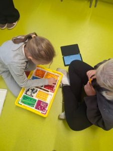 Dwie dziewczynki siedzą obok siebie na jasnozielonej podłodze. Przed nimi znajduje się otwarte pudełko LEGO z kolorowymi elementami, a obok leży tablet z wyświetlonym programem. Jedna dziewczynka wybiera klocek, druga trzyma pomarańczowy element w dłoni.