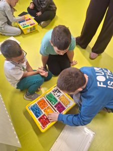 Trzech chłopców siedzi na jasnozielonej podłodze przy otwartym żółtym pudełku LEGO z kolorowymi klockami, wspólnie wybierając elementy. W tle inne dzieci pracują nad podobnym zestawem.