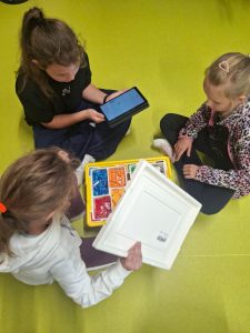 Trzy dziewczynki siedzą na podłodze przy otwartym pudełku LEGO. Jedna trzyma w rękach biały tablet, druga podnosi plastikową pokrywkę, a trzecia patrzy na pudełko z kolorowymi elementami.