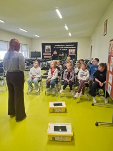 Grupa dzieci siedzi na krzesłach w sali i słucha stojącej przed nimi kobiety. Na podłodze leżą dwa zamknięte zestawy LEGO w żółtych pudełkach. W tle widać scenę z czarnym banerem „Wojewódzki konkurs plastyczno-recytatorski”