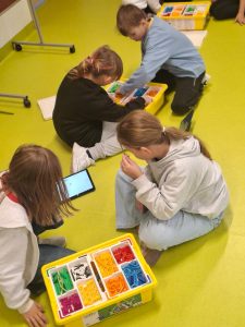 Czwórka dzieci siedzi na jasnozielonej podłodze w sali, pracując z zestawami LEGO Education w żółtych pudełkach. Jedno dziecko korzysta z tabletu, pozostałe wybierają klocki z przegródek.