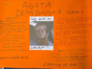 Plakat wyborczy na pomarańczowym tle z dużym, ręcznie napisanym czarnym tekstem. Na górze znajduje się napis: „AGATA DEMBIŃSKA KLASA 7”. W centralnej części plakatu wklejone jest zdjęcie dziewczyny w okularach, z podpisem: „TWÓJ GŁOS MA ZNACZENIE!!!”. Po lewej stronie znajduje się tekst: „Cześć Wam!!! Jestem uczennicą klasy 7. Interesuję się sportem, lubię czytać książki i słuchać muzyki. Jestem miła, grzeczna, sympatyczna, koleżeńska, uczynna i śmiała. Lubię pomagać starszym ludziom i dzieciom w różnych zadaniach i dziedzinach. Chciałabym włączyć się w pomoc na rzecz szkoły, klasy jak i uczniów. Lubię czynić dobro drugiemu człowiekowi. Nie lubię przemocy i agresji. Razem możemy więcej.” Po prawej stronie tekst w kilku akapitach: „Chciałabym, aby w naszej szkole było więcej zabaw (dyskotek) oraz zawodów sportowych. Mogłyby być prowadzone Dni Bez Plecaka, w których można by było zająć się sportem lub zabawą. Chciałabym, aby były Dni Zdrowego Żywienia, gdzie sami byśmy sobie robili jedzonko. W ciepłe dni mogłyby być lekcje na trawie na boisku szkolnym. Spróbuję zrobić jeden dzień w miesiącu bez sprawdzianów i kartkówek dla całej szkoły. Mam jeszcze wiele pomysłów, ale żeby się o nich przekonać musicie na mnie zagłosować, o co bardzo was proszę. Zagłosuj na mnie!!!”