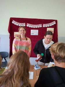 Dwie uczennice klasy ósmej stoją na tle tablicy zakrytej czerwonym materiałem, na której umieszczono zostało hasło: LUBIMY POMAGAĆ INNYM. Jedna z dziewczynek uśmiecha się szeroko a druga pochyla nad stołem stojącym przed nimi trzymając w dłoni ketchup.