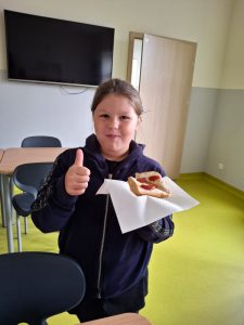 Uczennica stoi na szkolnej stołówce trzymając w lewej dłoni tost. W prawej dłoni ma wyciągnięty w górę kciuk.
