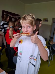 Uczeń stoi w szkolnej stołówce. Chłopiec je prawą dłonią tost, a lewą rękę ma podniesioną w górę z wyprostowanym kciukiem. W tle widać innych uczniów.
