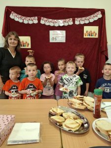 Uczniowie oddziału przedszkolnego stoją wraz z wychowawczynią na tle zawieszonego na ścianie czerwonego materiału ozdobionego napisem "LUBIMY POMAGAĆ INNYM". Dzieci i wychowawczyni trzymają w dłoniach tosty, a przed nimi na stole ułożone są półmiski z gotowymi kanapkami.
