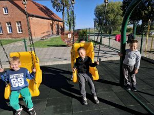 Trzech uczniów oddziału przedszkolnego korzysta z żółtych huśtawek na placu zabaw. Dzieci uśmiechają się, a w tle widoczne jest szkolne ogrodzenie i budynek.
