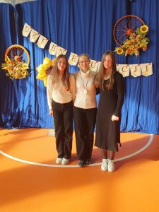Trzy osoby stoją na tle niebieskiej kotary ozdobionej girlandą z napisem „Dzień Edukacji”. Po bokach wiszą dekoracje z kwiatami i pomalowanymi kołami rowerowymi. Osoby uśmiechają się do zdjęcia, stojąc na pomarańczowej sali gimnastycznej.