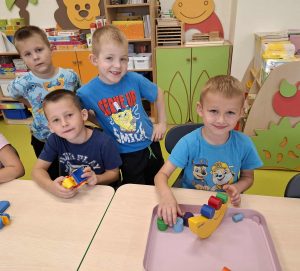 Grupa uczniów oddziału przedszkolnego bawi się przy stoliku w sali. Jeden z chłopców układa klocki na balansującej zabawce, a pozostali z uśmiechem obserwują zabawę.