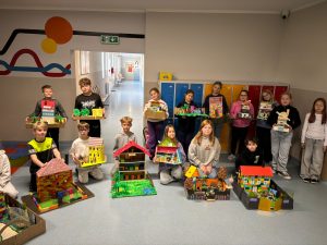 Grupa uczniów klasy IV stoi na szkolnym korytarzu i prezentuje wykonane makiety domów inspirowanych lekturą „Akademia Pana Kleksa”. Każdy uczeń trzyma własnoręcznie przygotowaną pracę z kartonu, klocków i materiałów plastycznych. Zdjęcie stanowi dokumentację projektu zrealizowanego na lekcji języka polskiego.
