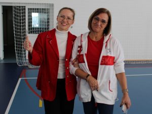 Dwie nauczycielki stojące razem na sali gimnastycznej. Jedna ubrana w czerwony żakiet pokazuje kciuk do góry, druga w białej sportowej bluzie z napisem „Polska” uśmiecha się i trzyma telefon w ręku.