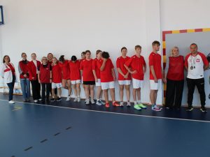 Grupa kilkunastu osób stojących w rzędzie przy ścianie sali gimnastycznej. Uczestnicy mają czerwone koszulki i białe spodenki sportowe, obok nich stoją trenerzy i opiekunowie w czerwonych strojach.