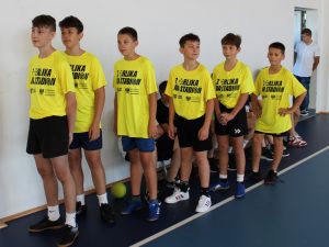 Grupa sześciu chłopców w żółtych koszulkach sportowych z napisem „Z Orlika na Stadion”, stojących w rzędzie na sali gimnastycznej. Chłopcy mają krótkie spodenki i sportowe buty, obok nich widać piłkę.