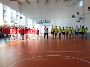 Dwie drużyny sportowe ustawione w rzędach na sali gimnastycznej – po lewej w czerwonych koszulkach, po prawej w żółtych – a między nimi stoi trener.